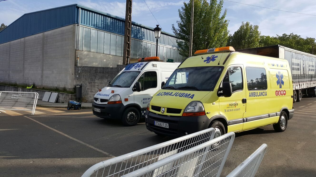 Dos ambulancias de Ambulancias do Nordés estacionadas junto a una nave industrial, con vallas de seguridad y un camión al fondo